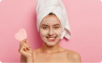 Gorgeous gentle woman with pleasant smile bites lower lip has granules sea salt applied around face holds cosmetic sponge wiping complexion wears towel after bath has well cared body